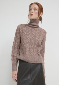 Eine rothaarige Frau trägt einen braunen, strukturierten Rollkragenpullover und einen schwarzen Lederrock. Sie schaut leicht nach oben und zur Seite.