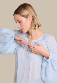 Lys blå, gjennomsiktig bluse med puffede ermer, en samlet halslinje og en nøkkelhulldetalj. Myk tekstur med flytende stoff.