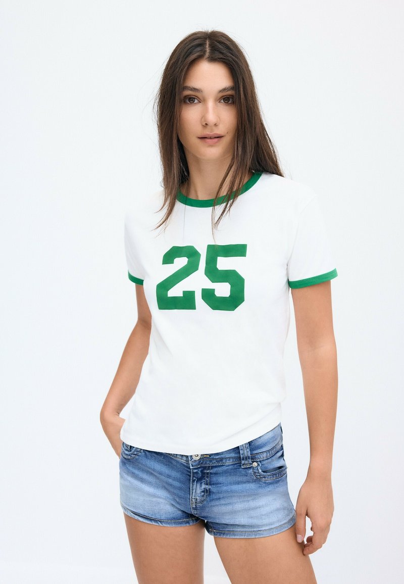 Jeune femme debout portant un t-shirt blanc avec le numéro 25 vert et une bordure verte, associé à un short en denim bleu sur un fond blanc uni.