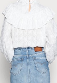 Vit blus med volangkrage och volangärmar, mönstrad textilstruktur. Bärs över en ljusblå midjebyxa av denim med bakfickor.