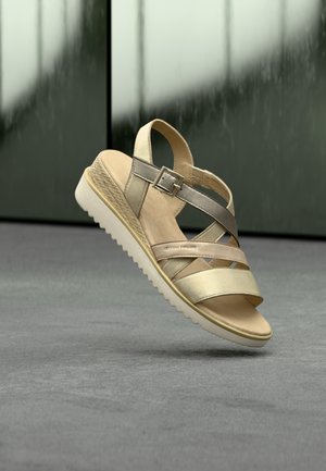 Bézs és fémes pántos szandál csattal, texturált ékarással és fehér, bordázott talppal, amely szürke padló és sötét háttér felett lebeg.