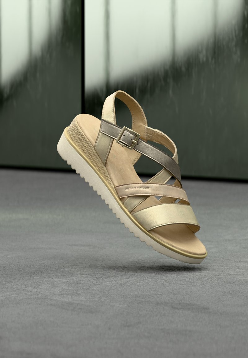 Μπεζ και μεταλλικό σανδάλι με λουράκια και αγκράφα, με τακούνι πλατφόρμας με υφή και λευκή αυλακωτή σόλα, αιωρούμενο πάνω από γκρι πάτωμα και σκοτεινό φόντο.