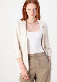 Svetlobežna lanena blazer s pokrobnimi rokavi in notch ovratnikom, v kombinaciji z belim topom s kvadratnim izrezom in bež črtastimi hlačami.
