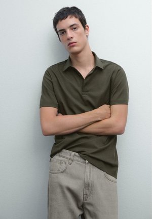 T-shirt polo vert olive avec un col, des manches courtes et un tissu texturé. Associé à un jean droit gris clair. Design simple.