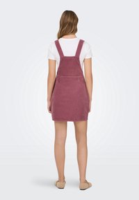 Femme debout, de dos, portant une chemise blanche à manches courtes, une robe salopette en velours côtelé rose et des chaussures beiges à enfiler sur un fond uni.