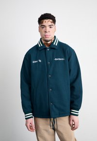 Mørk teal jakke med hvit brodering, grønn og hvit stripete krage, knappefront og side lommer. Stoffet ser teksturert og solid ut.