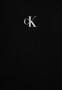 T-shirt preta de algodão com um logótipo cK branco ao centro. O tecido é liso, com uma textura consistente e sem padrões ou acentos visíveis.