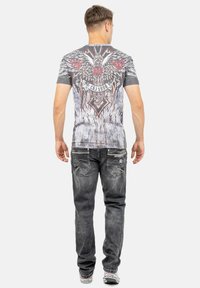 Grijze T-shirt met een grafisch ontwerp met rode accenten, vleugels en sierlijke details op de achterkant, gecombineerd met vervaagde zwarte jeans.