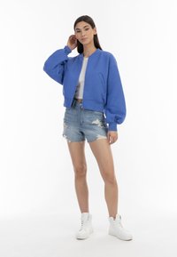 Giacca bomber imbottita blu con polsini e orlo a costine, abbinata a pantaloni corti in denim strappati e sneakers alte bianche.