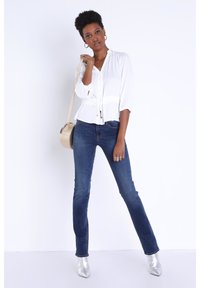 Weiße Bluse mit Taillenknoten, Dreiviertelärmel. Dunkelblaue Schlagjeans. Silberne spitze Ankle Boots; goldene runde Handtasche.