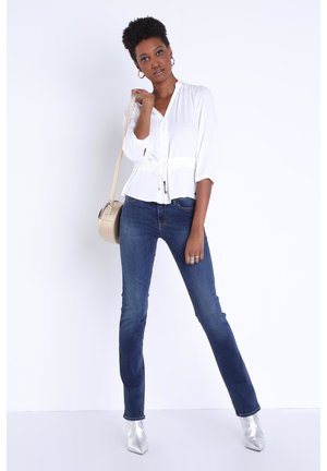 Weiße Bluse mit Taillenknoten, Dreiviertelärmel. Dunkelblaue Schlagjeans. Silberne spitze Ankle Boots; goldene runde Handtasche.