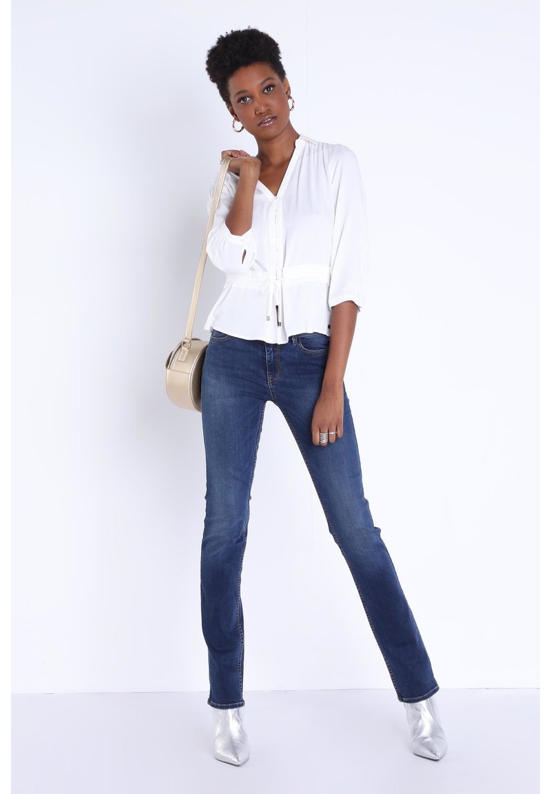Weiße Bluse mit Taillenknoten, Dreiviertelärmel. Dunkelblaue Schlagjeans. Silberne spitze Ankle Boots; goldene runde Handtasche.