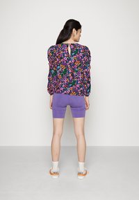 Blusa floral multicolorida com mangas bufantes, detalhe de buraco na parte de trás, combinada com calções de ciclismo roxos e ténis brancos com acentos laranja.