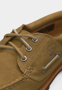 Chaussure bateau en daim beige avec des lacets en cuir passant à travers des œillets en métal et le logo Timberland en forme d'arbre embossé sur le côté.