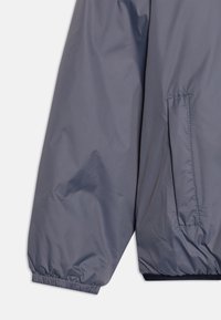 Giacca leggera grigia con polsini a coste, tasca con zip e una texture di tessuto liscia con sottili righe orizzontali.