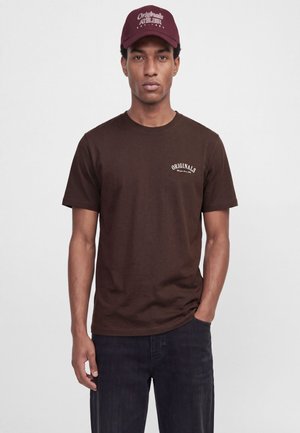 Homme portant un t-shirt marron foncé avec le texte "Originals" sur la poitrine et une casquette bordeaux avec texte blanc, debout devant un fond blanc.