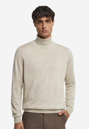Un jeune homme portant un pull turtleneck beige et un pantalon marron, se tenant avec une main dans la poche contre un fond blanc.