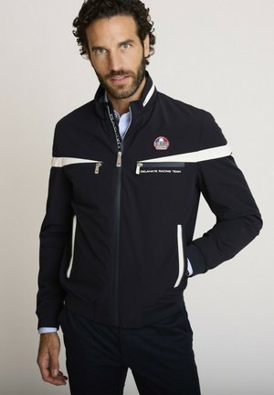 Homme portant une veste bleu marine foncé avec des rayures blanches et le logo Delahaye Racing Team, une main dans la poche, sur un fond clair.