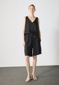 Svart satinlinne utan ärmar med V-ringning, parat med plisserade svarta shorts. Modellen bär guldiga klackar med remmar. Enkelt och elegant design.