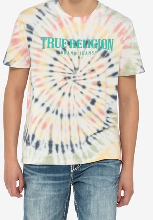Osoba ubrana w wielokolorową koszulkę tie-dye z napisem "TRUE RELIGION BRAND JEANS" oraz niebieskie dżinsy na jednolitym tle.