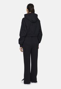 Geknipte zwarte hoodie met een brede capuchon, losse mouwen en een elastische taille; bijpassende zwarte wijde broek; gladde textuur. Witte sneakers worden gedragen.