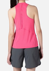 Žena v růžovém bezrukávovém sportovním topu s mřížkovaným detailem na zadní straně a tmavě šedých šortkách, stojící zády k fotoaparátu na bílém pozadí.