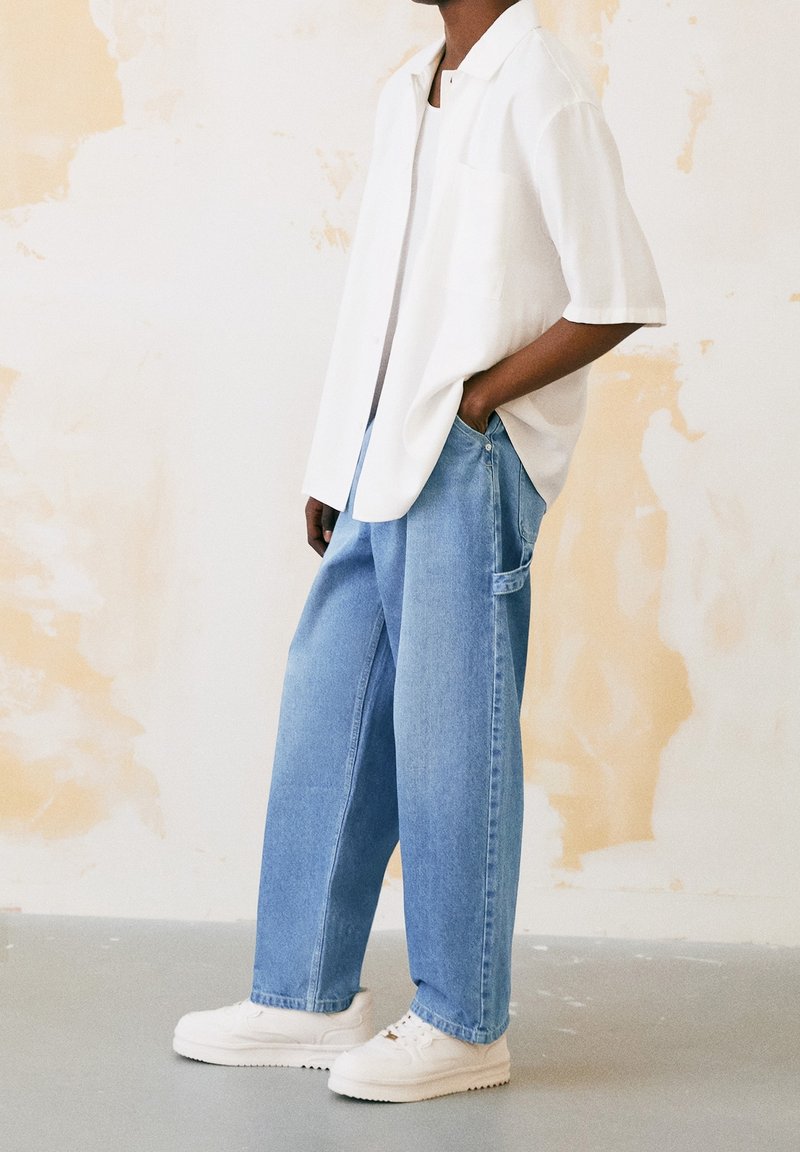 Osoba w luźnych jasnoniebieskich dżinsach, białych trampkach i krótkim białym koszuli z jedną ręką w kieszeni, stojąca przy beżowej ścianie.