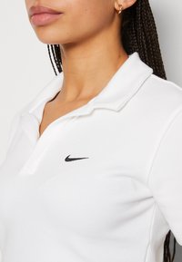 Vit bomullspolo med en svart Nike-logotyp på vänster bröst, med en klassisk krage och korta ärmar. Slät textur.