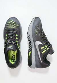 Baskets de sport noires et grises avec une tige texturée, des accents vert fluo et une semelle rembourrée. Présente un label de soutien dynamique sur le côté.