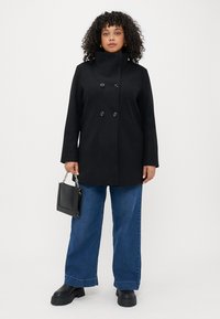 Cappotto nero doppiopetto con colletto alto, con grandi bottoni. Abbinato a jeans in denim a gamba larga e stivali neri robusti.