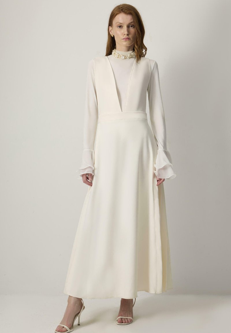 Lang witte jurk met een diepe V-hals, lange doorzichtige mouwen, een geraffineerde taille en een vloeiende rok. Bevat een gestructureerde kraag en delicate manchetjes.