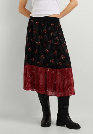 Frau trägt einen schwarzen und roten Midirock mit Blumenmuster und Rippenstruktur, kombiniert mit schwarzen kniehohen Lederstiefeln und einem weißen Oberteil.