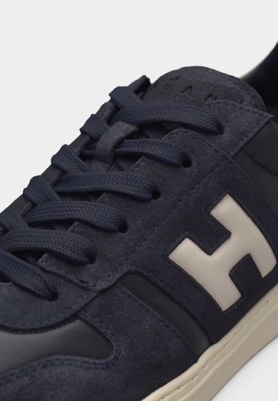 Κοντινή λήψη σνίκερ από ναυτικό μπλε σουέντ και δέρμα με κορδόνια του ίδιου χρώματος και μεγάλο λευκό λογότυπο H στο π λάι πάνω από λευκή σόλα.