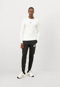Langermet hvit sportslig skjorte med svart Nike-logo, kombinert med svarte joggebukser med hvit grafikk, og hvite joggesko med røde detaljer.