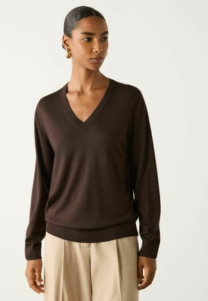 Kvinde iført en mørkebrun V-hals sweater og beige bukser med brede ben, med håret bundet tilbage og gyldne hoop øreringe, kigger let mod højre.