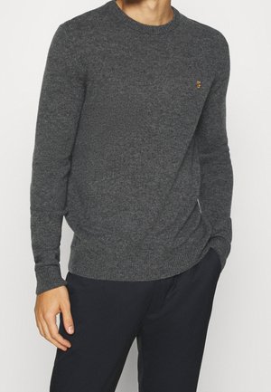 Mężczyzna ubrany w dopasowany, ciemnoszary sweter z długim rękawem typu crewneck oraz czarne spodnie, jedna ręka w kieszeni, na jednolitym tle.