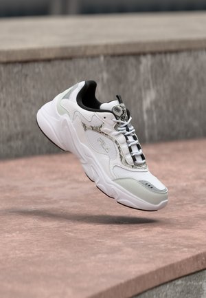 Weißer und grauer FILA-Sneaker mit strukturierten Details und Schnürsenkeln, schwebend über einer rosa-steinernen Oberfläche in der Nähe von Betontreppen.