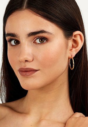 Goldene Creolen mit einem gedrehten Design, glänzender Oberfläche und einem kleinen Verschluss, getragen von einer Person mit glattem, dunklem Haar.