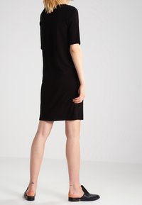 Czarna sukienka z krótkim rękawem, prostym krojem, do kolan, o minimalnej teksturze. Noszona z czarnymi butami wsuwanymi. Skóra widoczna na nogach.