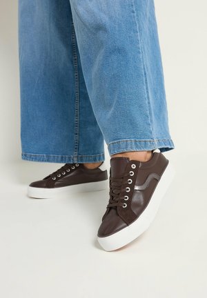 Personne portant un jean large bleu et des baskets marron foncé avec des semelles blanches épaisses, debout sur un fond clair.