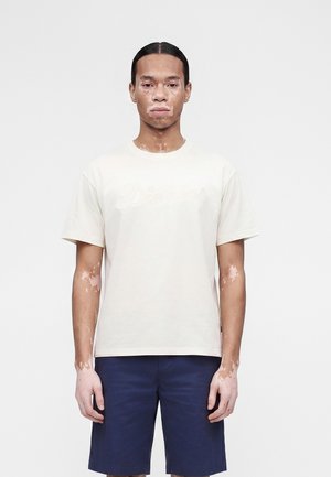 Νεαρό άτομο με λεύκη φοράει ένα λευκό κοντομάνικο πουκάμισο και σκούρο μπλε σορτς, όρθιο μπροστά σε απλό ανοιχτό φόντο.