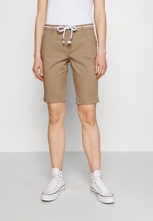 Person iført beige knælange shorts med hvid snor, hvid ærmeløs top og hvide høje sneakers, der står på en grå gulv.