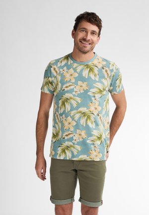 Korte ærmer, T-shirt i bomuld med en lys blå baggrund, der har et tropisk blomstermønster i gult og grønt. Sammen med op rullede khaki shorts.