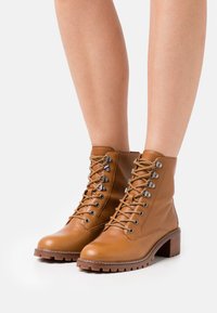 Brązowe skórzane botki za kostkę z okrągłym noskiem, sznurowanym zapięciem, metalowymi oczkami i teksturowaną podeszwą. Posiadają drewniany obcas w kształcie bloku.