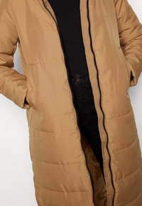 Personne portant une doudoune beige avec une fermeture éclair noire, les mains dans les poches, un chemisier noir et un pantalon noir taille haute avec des boutons visibles.