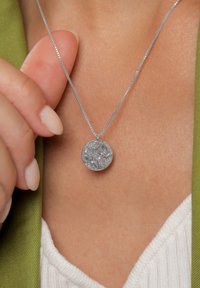 Srebrny naszyjnik z zawieszką o teksturowanej, okrągłej formie, zawieszony na delikatnym łańcuszku typu wężyk, spoczywający na skórze na tle zielonego ubrania.