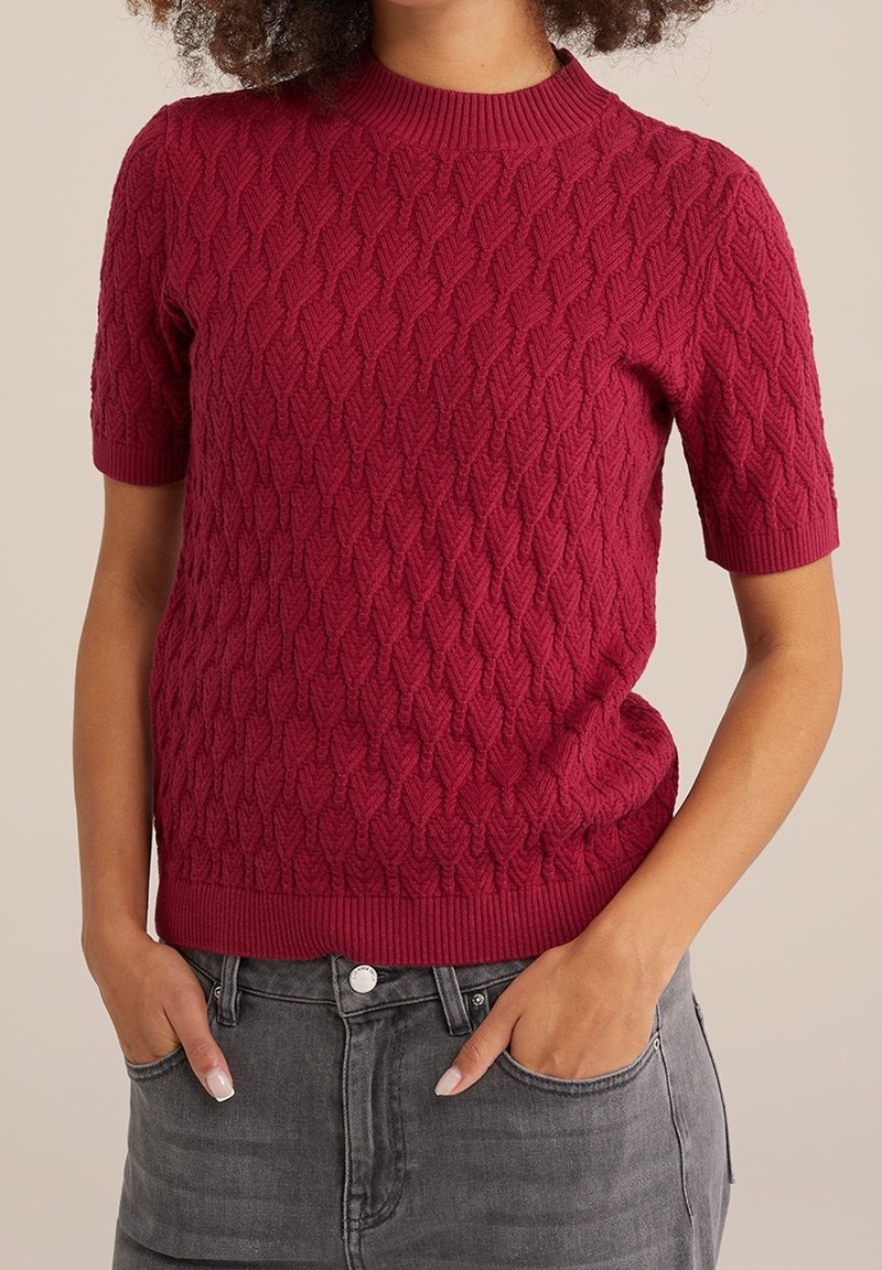Roter kurzärmliger Strickpullover mit strukturiertem Kabelmuster, mit hohem geripptem Kragen und geripptem Saum, kombiniert mit grauen Jeans.