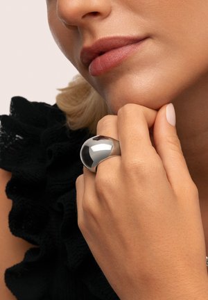 Bague métallique argentée avec une forme de dôme lisse et arrondi. Main posée sur le menton montrant la texture de la peau et un léger vernis à ongles sur les doigts.