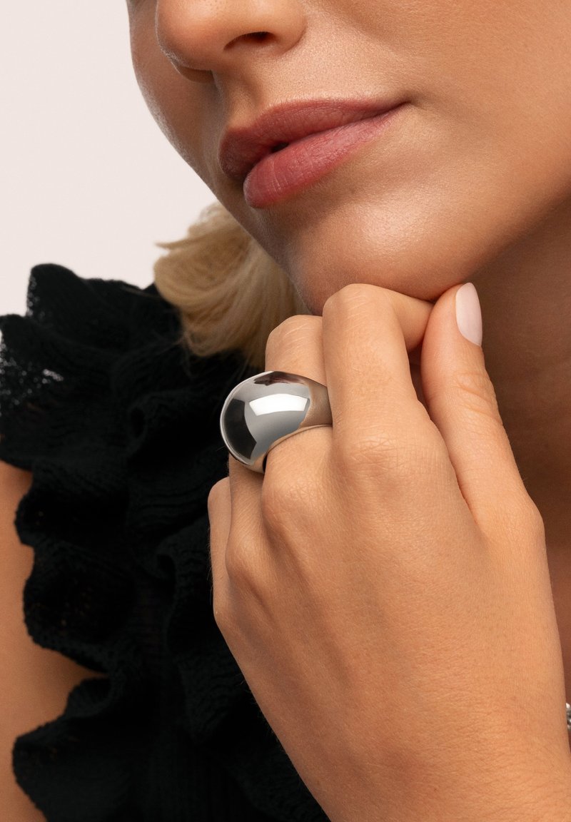 Svensksilver metallic ring med en slät, rund kupolform. Handen vilar på hakan och visar hudens struktur samt ett ljust nagellack på fingrarna.