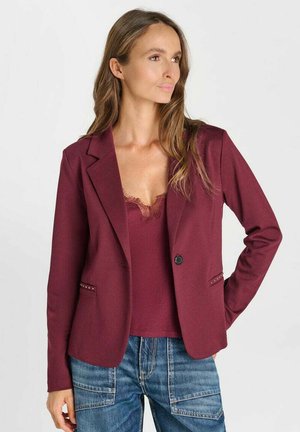 Vrouw draagt een donkerrode blazer en een bijpassende top met kanten rand, gecombineerd met blauwe spijkerbroek, kijkt opzij met een hand op haar heup.